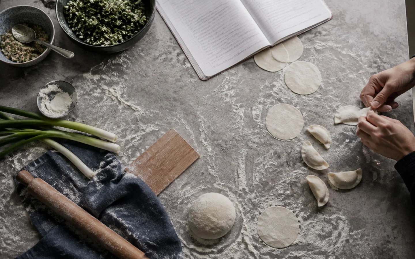 Hands folding dumplings beside a handwritten recipe notebook
